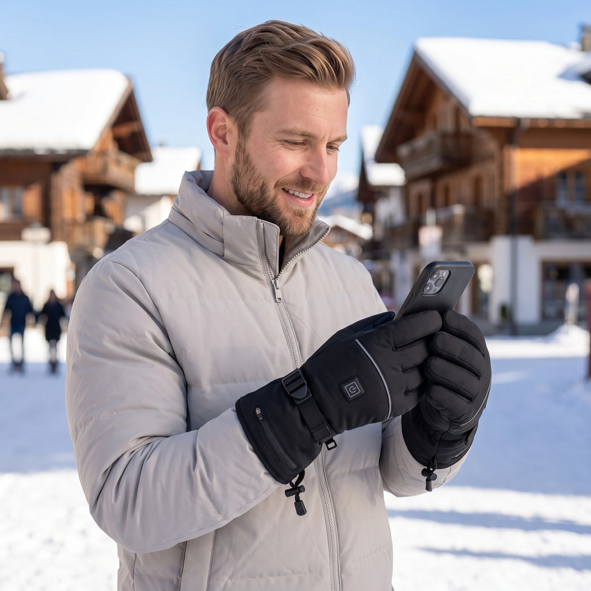 Homme souriant qui consulte son téléphone dans un village enneigé, gants chauffants aux mains